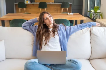 Young woman relaxing on a sofa with a laptop, stretching and smiling while working from home in a bright modern living room, feeling comfortable and content. © simona