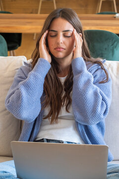 Young woman with headache rubbing temples while working on laptop at home, feeling stressed and tired