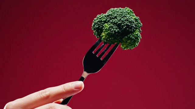 Hand holding a fork with a piece of broccoli against a vibrant red background in a studio shot