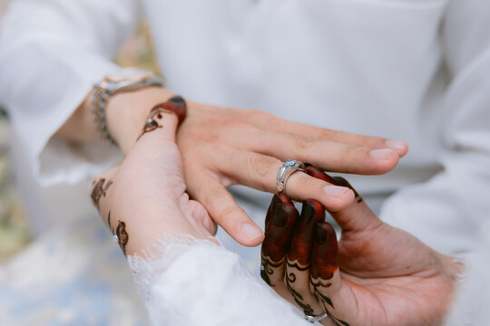 Close-up image of a bride's henna-painted hand slipping a silver ring with a blue gemstone onto the groom's finger during their wedding ceremony.