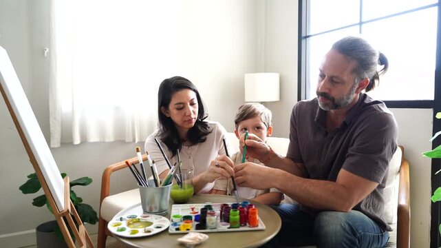 The family&mdash;mother, father, and son&mdash;are learning to paint with watercolors. The mother is teaching, the parents are teaching their son, and they are chatting as a hobby in the living room at home.