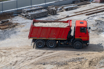 a bright red dump truck fully loaded with sand driving across the dirt, with stacked concrete slabs and other building materials visible in the background. © Andrey