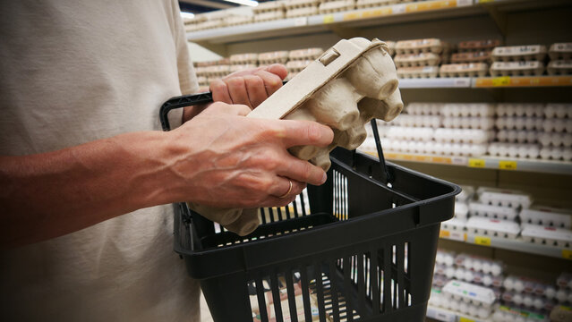 Close-up of a customer's hand putting a carton of eggs into a grocery basket in a supermarket egg aisle. Retail daily, grocery shopping and household lifestyle concept
