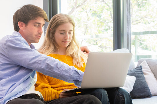 Young couple sitting on a couch at home using a laptop together, sharing ideas and online browsing in a bright living room, relaxed and focused while working, studying, or shopping from home.