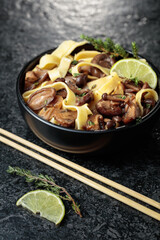 Fototapeta premium Shimeji and shiitake mushrooms stir-fried with soy sauce, garlic and thyme, served with noodles and lime slice in a black ceramic bowl.