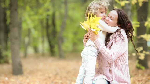 A happy young family with a child is having fun in the autumn park. Mother and daughter is hugging outdoors against blurred autumn leaves background. Slow motion