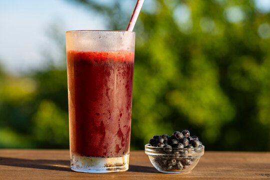 Refreshing blueberry smoothie in misty glass with fresh berries, closeup. Cold organic berry smoothie or juice in frosted glass with straw next to bowl of fresh blueberries on wooden table outdoors