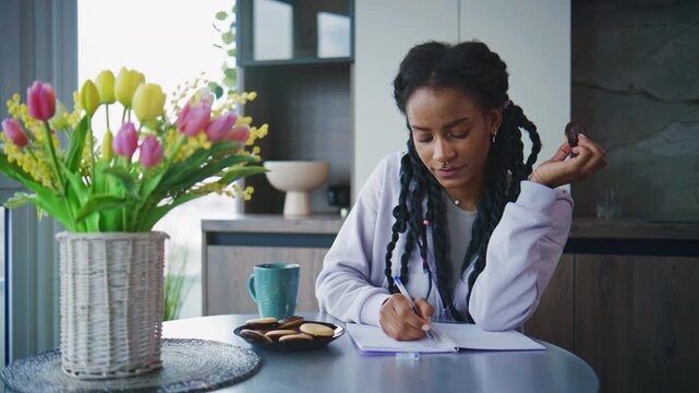 Woman writing in a notebook while sitting at a table with flowers and snacks in a bright indoor setting during the day