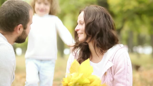 A happy young family with a child is having fun in the autumn park. Mother, father and daughter is hugging outdoors against blurred autumn leaves background. Slow motion