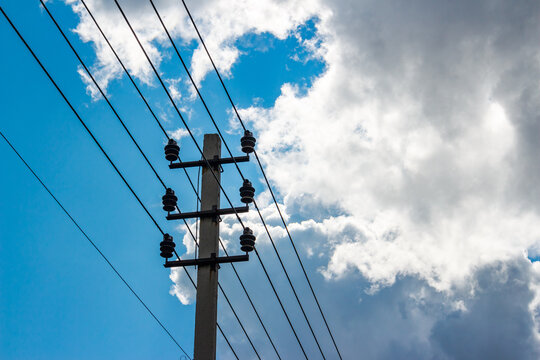 Concrete utility pole with high voltage power lines stretching across a bright blue sky with dramatic white fluffy clouds, low angle shot of electrical infrastructure against daylight