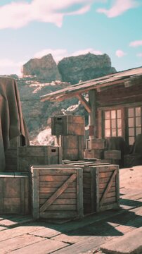 Wooden crates are stacked near a building in a dry desert landscape. The scene shows a clear sky and rock formations in the background. Sunlight casts shadows on the ground.