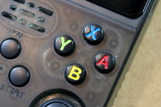 Color-coded Y, X, A and B buttons on a black retro handheld emulator console, extreme close-up of generic gaming controls with start key, speaker grille and dusty plastic surface, no people
