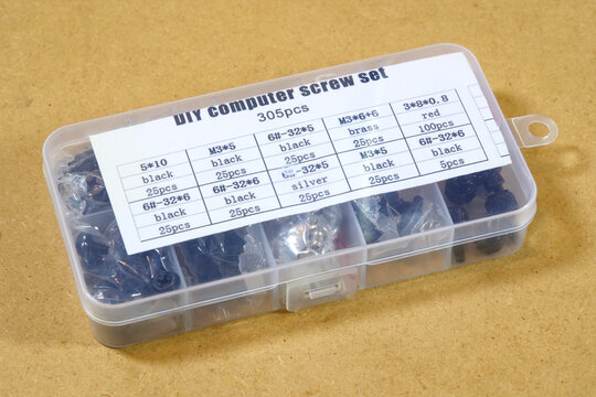 Clear plastic organizer box containing a 305-piece computer screw set with black, silver, brass fasteners on a brown workbench, close-up high angle view, DIY electronics repair supplies