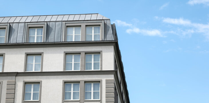 Modern gray multi-story building facade with white windows and metal mansard roof beneath a blue sky, wide panoramic urban architecture exterior with large copy space and no people