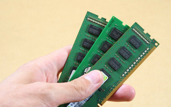 Hand holding three green desktop RAM modules with black chips and gold contacts, close-up of computer hardware upgrade, component shortage, limited supply and rising technology prices concept