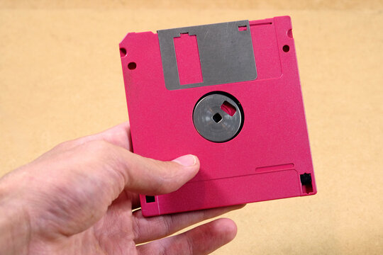 Hand holding a bright pink 3.5-inch floppy disk above a brown surface, close-up of retro computer data storage, save icon, obsolete magnetic media and 1980s technology nostalgia concept