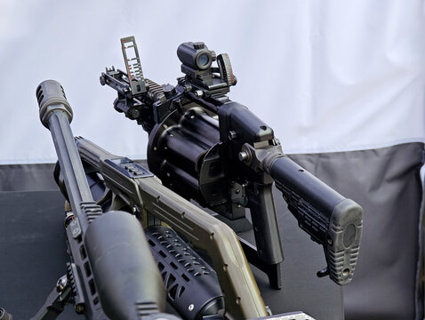 Modern rotary grenade launcher with drum magazine, optical sight and adjustable ladder sight displayed among other firearms, close-up rear view of tactical military weapon hardware indoors, no people