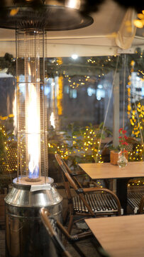 Tall gas patio heater with visible flame beside empty restaurant tables and woven chairs in an enclosed outdoor garden at night, warm string light bokeh, cozy dining terrace, vertical no people scene