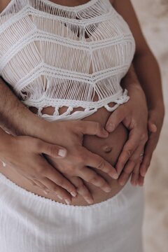 Close-up view of couple hands forming heart shape on pregnant belly with white macrame top