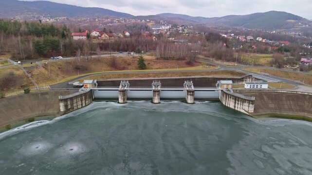 In Świnna Poręba, Poland, the Zapora Świnna Poręba dam spans the frozen Mucharskie reservoir amid winter forests and mountains, viewed by drone as part of regional water management infrastructure.