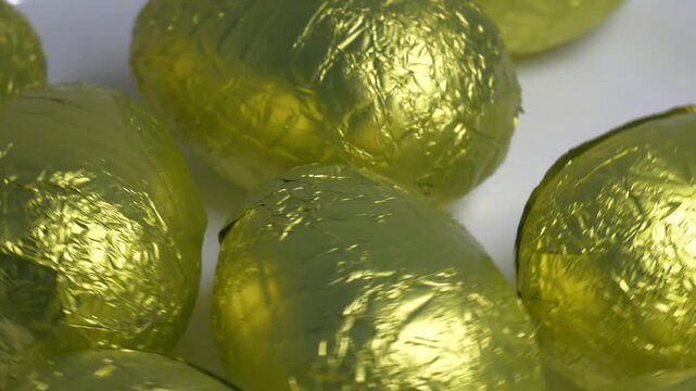 Close-up of chocolate Easter eggs wrapped in shiny golden foil rotating on a white plate, highlighting their textured metallic surface under bright lighting.