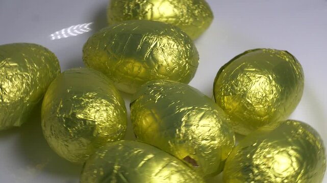 Close-up of chocolate Easter eggs wrapped in shiny golden foil rotating on a white plate, highlighting their textured metallic surface under bright lighting.