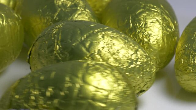 Close-up of chocolate Easter eggs wrapped in shiny golden foil rotating on a white plate, highlighting their textured metallic surface under bright lighting.