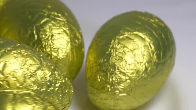 Close-up of chocolate Easter eggs wrapped in shiny golden foil rotating on a white plate, highlighting their textured metallic surface under bright lighting.