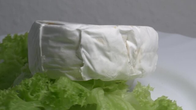 Close-up of a piece of Brie cheese on fresh green lettuce leaves rotating on a white plate, showcasing its soft texture and white rind under studio lighting.