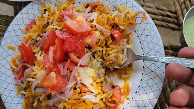 Person eating spicy chicken biryani topped with fresh tomatoes and onions
