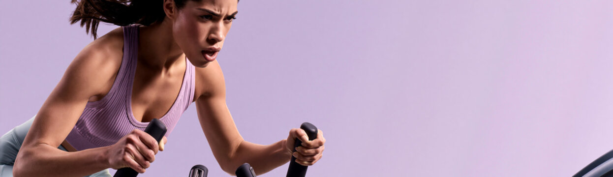 Athletic young woman doing intense cardio workout on exercise machine. Focused female athlete in purple sports bra on matching background. Fitness training concept with copy space for text