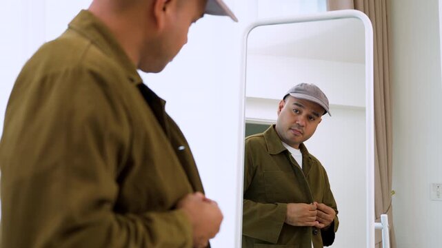 Smiling Asian man in a baseball cap looking in a mirror, adjusting his brown casual shirt jacket. Concept of morning preparation, urban street fashion and everyday confidence.