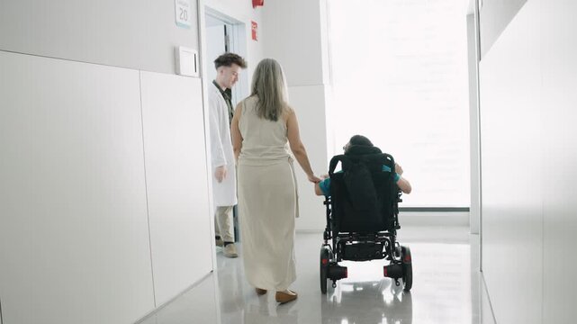 Woman pushing a young man with a physical disability in a wheelchair along a modern hospital hallway. He suffers from spina bifida