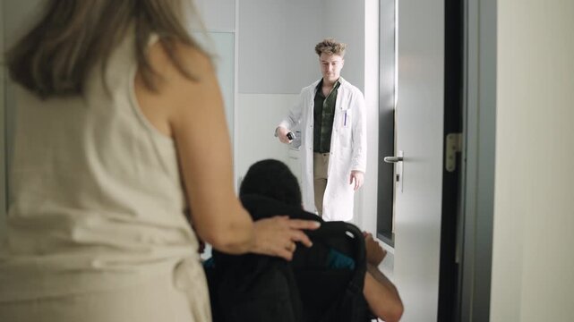 Young man with spina bifida in a wheelchair, accompanied by his mother, entering a doctor's office for a medical appointment at the hospital