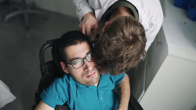 Male doctor using an otoscope to check the ear of a young adult man with spina bifida sitting in a wheelchair during a medical checkup