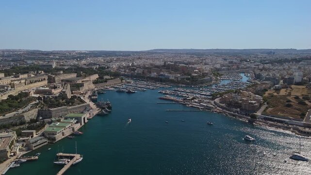 Over Marsamxett Harbour in Malta, an aerial view captures yachts and boats moored along the urban waterfront, set against historic fortifications and Mediterranean architecture.