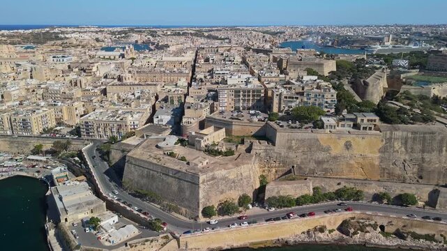 In Floriana overlooking Valletta and Marsamxett Harbour, the historic Maltese cityscape with stone fortifications and waterfront buildings spreads across the bay in a drone view.