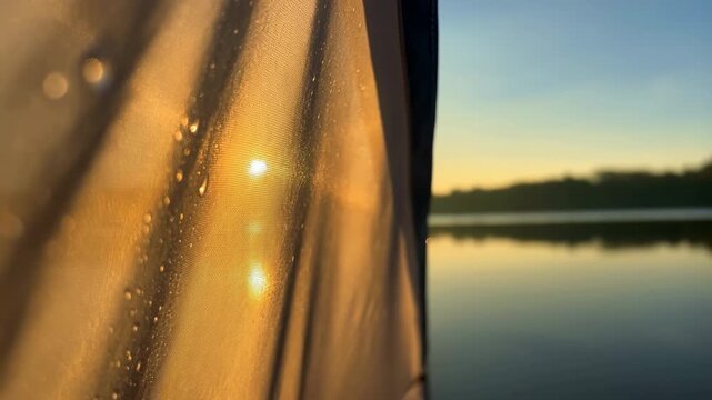 Backlit translucent fabric panel beside lakeshore at sunrise, shifting from cool blue tones toward warm golden highlights with woven texture, stitched hem, droplets, soft horizon glow, and calm