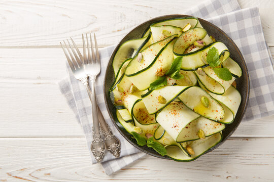 Zucchini Carpaccio is a raw courgettes salad with with pistachios, chili pepper and mint close-up in a plate on the table. Horizontal top view from above