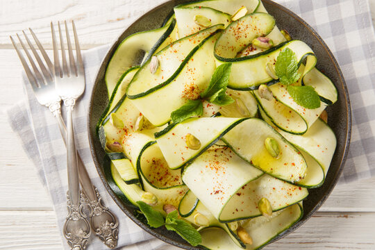 Zucchini carpaccio is a fresh, crisp, and low-calorie appetizer topped with pistachios, chili pepper, and mint. A close-up of this appetizer on a plate. Horizontal top view from above