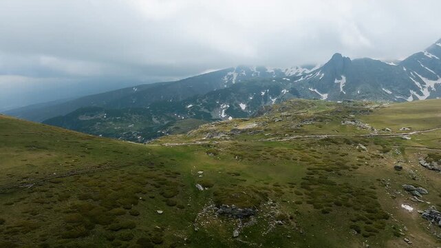 Seven Rila Lakes national park epic aerial drone view green grassy slope mountain peaks with patches of snow cloudy sky hiking destination summer travel adventure Bulgaria landscape.