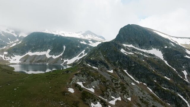 Seven Rila Lakes high mountain glacial lake peak with snow patches grassy ridge hiking trail panoramic drone view summer outdoor adventure destination Rila national park Bulgaria landscape.