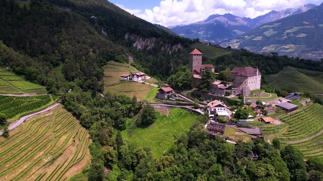 Castel Tirolo South Tyrol Italy 4K Drone. Pull back aerial flight from medieval castle with high altitude view of green vineyard terraces. Alpine mountain landscape scenery b roll video asset