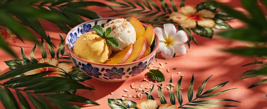 Scoops of peach and coconut ice cream decorated with mint and toasted coconut flakes in patterned bowl set on bright tropical floral tablecloth background with frangipani blossoms and palm foliage.