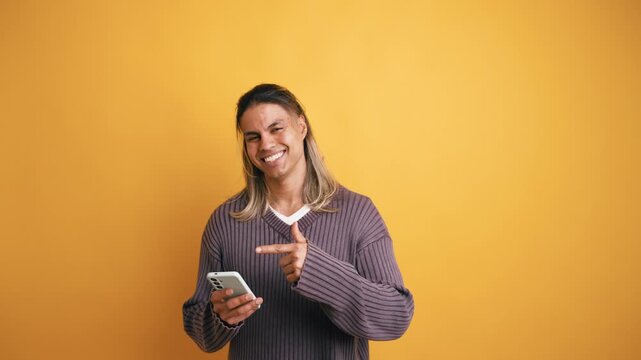Cheerful young Latino man smiling and pointing at his mobile phone screen. Happy guy enjoying a new app or service on a yellow background