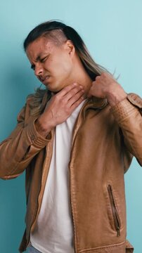 Young man with long hair wearing a leather jacket experiencing severe throat pain, touching his neck with a pained expression in a studio. Vertical