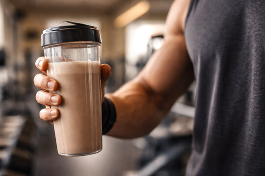 man holding shaker with protein drink in gym. Athlete drinking chocolate shake after workout. Nutrition and supplementation for muscle recovery. Healthy lifestyle and fitness concept