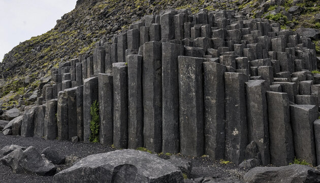 Dark grey basalt columns of a volcanic rock formation in iceland with a rocky ground and green moss growing on the side of the stone pillars