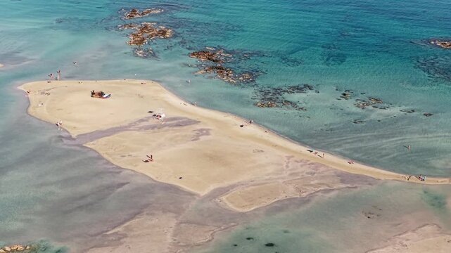 Aerial drone view of a broad sandy islet in the shallow turquoise lagoon fringed by rocky reefs, Elafonissi, Greece