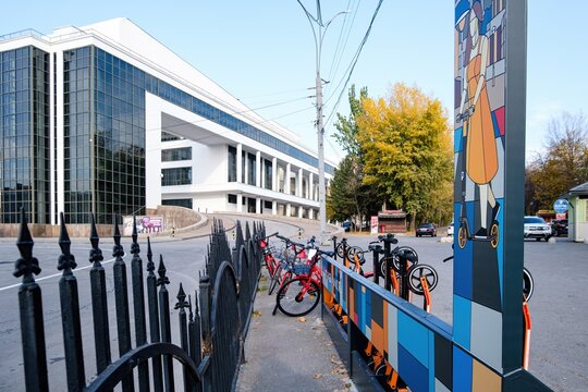 Rostov-on-Don, Russia - October 23, 2019: Urban micromobility hub with bright orange shared scooters and a red bicycle parked near a black charging kiosk, set against the Maxim Gorky Theater